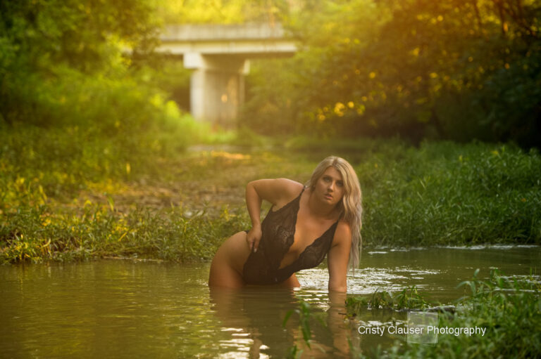 A woman in a black lace bodysuit poses knee-deep in a shallow, green creek surrounded by lush vegetation, with a bridge in the background and soft sunlight filtering through the trees. Cristy Clauser Photography