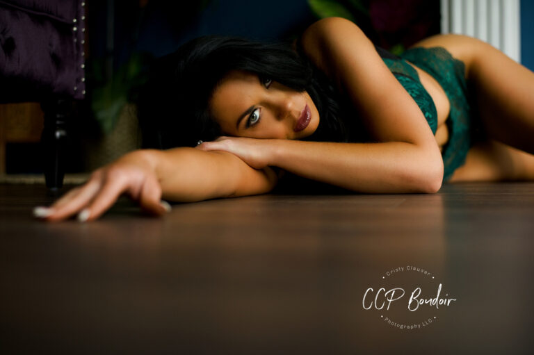 A woman in green lingerie lies on a wooden floor, gazing at the camera with one arm extended forward. The background is softly blurred, and the CCP Boudoir Photography logo appears in the lower right corner. Cristy Clauser Photography