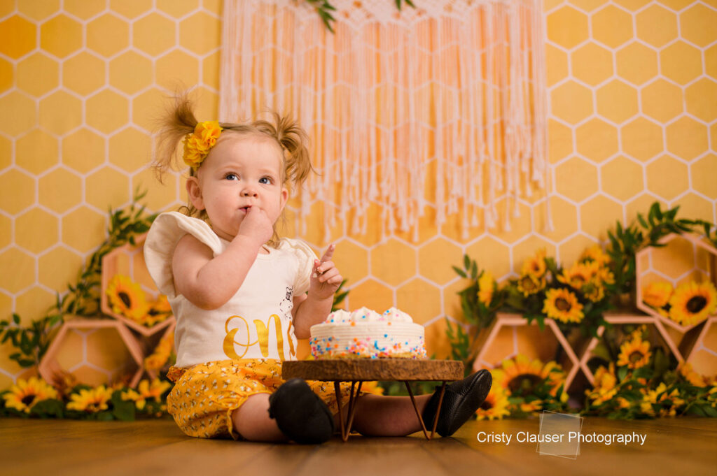 A toddler with pigtails and a yellow bow sits in front of a small cake with white frosting and sprinkles. The background is decorated with a honeycomb pattern and sunflowers. The child is wearing a white and yellow outfit.