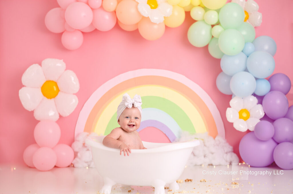 A smiling baby with a white bow sits in a small white bathtub. The background features pastel-colored balloon decorations, including a rainbow and flowers, set against a pink backdrop. Colorful petals and balloons surround the scene.