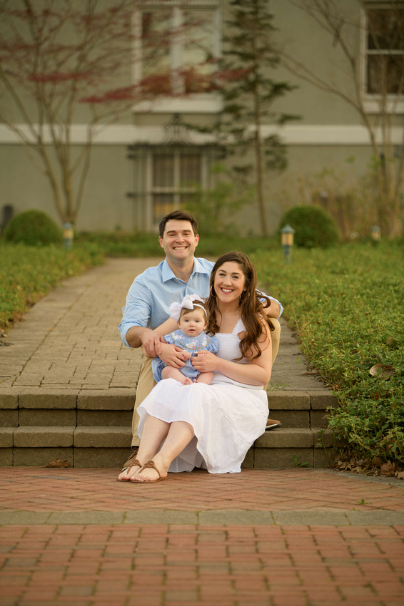 A smiling family of three—man, woman, and baby—sit together on outdoor stone steps. The parents wear light blue and white clothes; the baby wears a blue dress with a headband. Greenery and a house are in the background. Cristy Clauser Photography