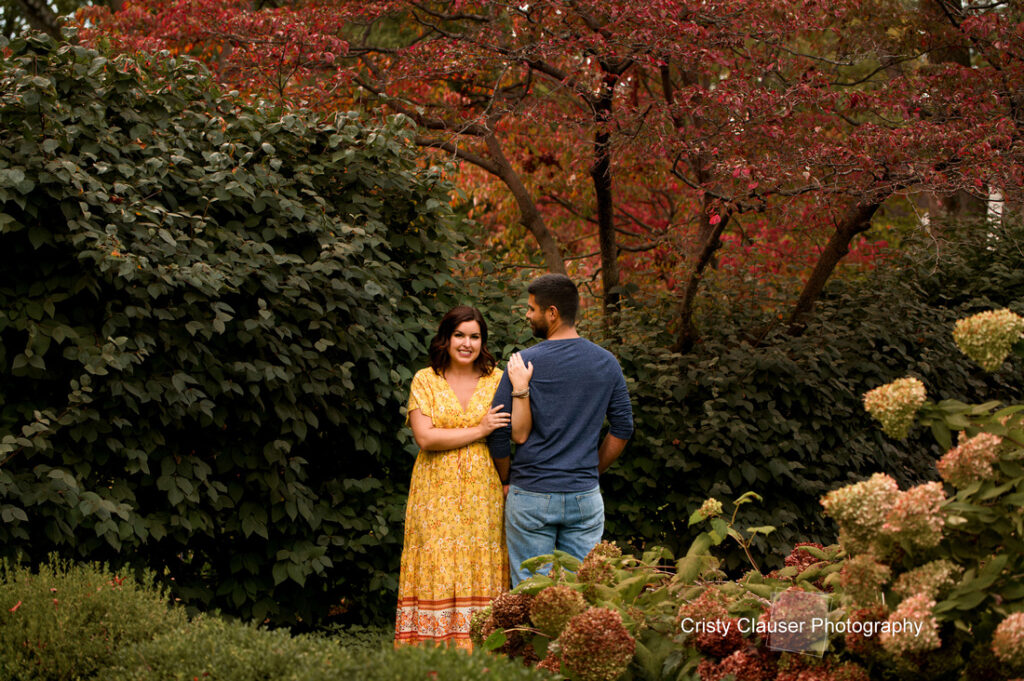 A woman in a yellow dress smiles while standing beside a man in a blue shirt in a lush garden with green bushes and red-leaved trees. The man faces away, and the woman holds his arm. Cristy Clauser Photography