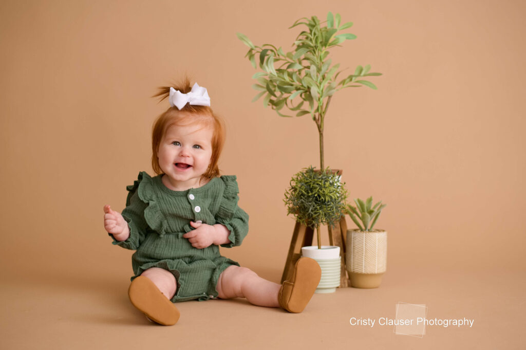 A smiling toddler with red hair, wearing a green outfit and white bow, sits on a beige floor next to three potted plants with a tan background. The photo is credited to Cristy Clauser Photography. Cristy Clauser Photography