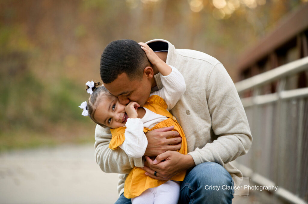 A father lovingly hugs and kisses his young daughter, who is smiling and wearing a yellow dress with white bows in her hair. They are sitting outdoors by a railing with a blurred natural background. Cristy Clauser Photography