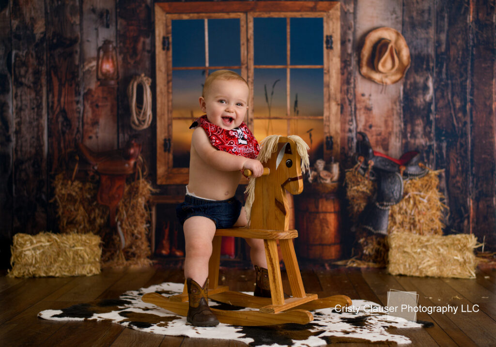 A baby wearing a red bandana, denim shorts, and cowboy boots smiles while sitting on a wooden rocking horse. The scene is set in a rustic room with straw bales, cowboy hats, a window showing a sunset, and wooden walls.