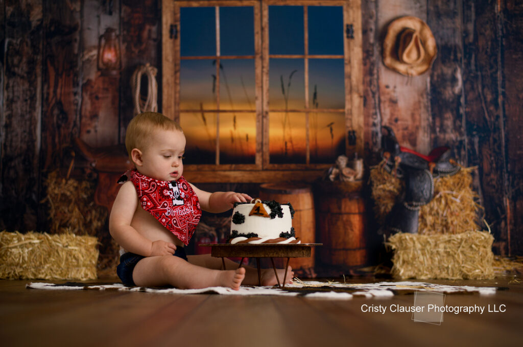 A baby wearing a red bandana sits on a wooden floor, touching a cake with a western theme. The rustic background features hay bales, a window with a sunset view, and cowboy decor.