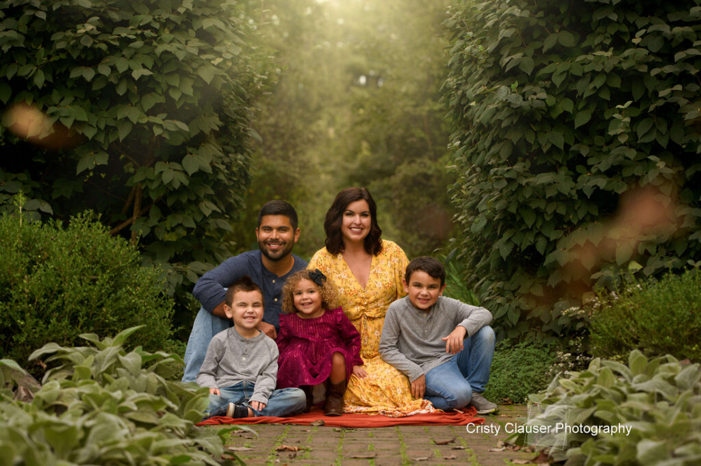 A family of five poses outdoors on a leafy path. The parents sit with their three young children—two boys and one girl—smiling at the camera, surrounded by lush green foliage. The mother wears a yellow dress. Cristy Clauser Photography