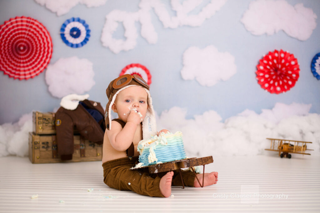 A baby is sitting on the floor eating a cake with blue and white frosting. The baby wears aviator goggles and a pilot hat. There's a toy airplane, brown jacket, and suitcase nearby, with cloud and target decorations in the background.