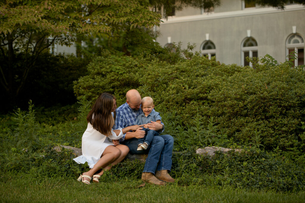 A family of three sits on a stone bench surrounded by greenery. The parents, a man and woman, smile at their young child, who is sitting between them, all appearing happy and relaxed outdoors. Cristy Clauser Photography