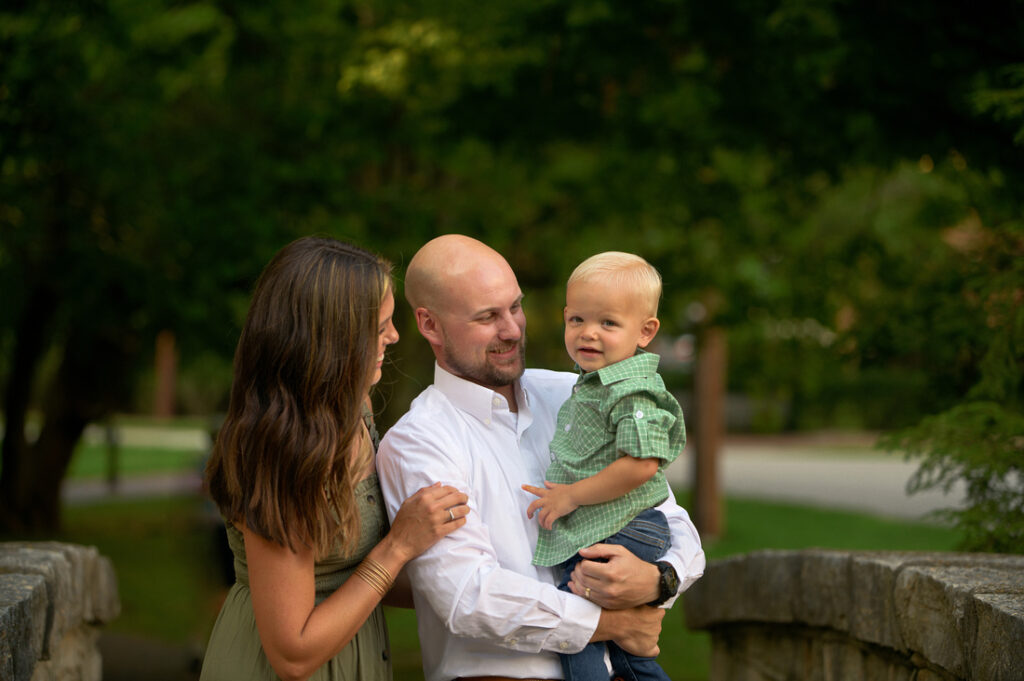 A woman and a man stand outdoors on a stone bridge, smiling at a young blond boy in the man's arms. The background is lush and green with trees. Cristy Clauser Photography